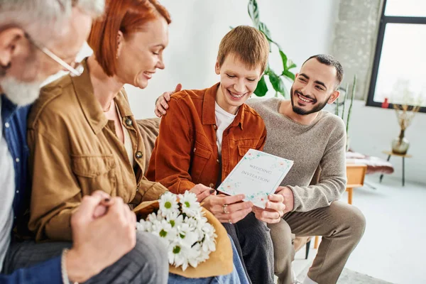 Cheerful gay man holding wedding invitation near boyfriend and happy mother with flowers in living room — Stock Photo