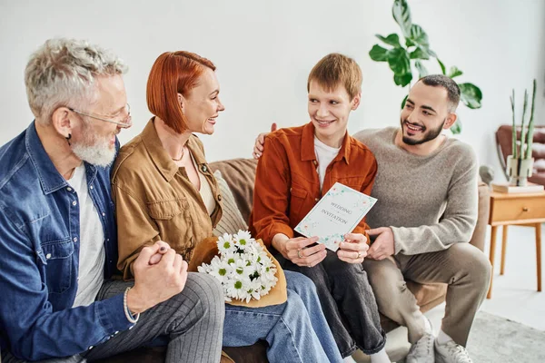 Pleased gay man showing wedding invitation to parents sitting on couch with flowers — Stock Photo