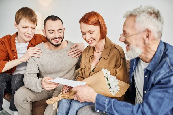 Overjoyed parents holding flowers and wedding invitation near son with gay partner at home — Stock Photo