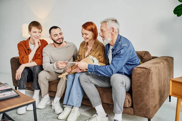 Cheerful gay man giving wedding invitation to parents while sitting on couch in living room — Stock Photo