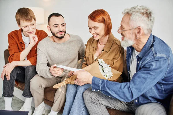Smiling gay man giving wedding invitation to parents near boyfriend on sofa in living room — Stock Photo