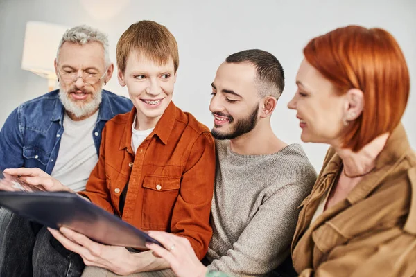 Радісний гей чоловік посміхається біля хлопця і батьків, тримаючи фотоальбом у вітальні — стокове фото
