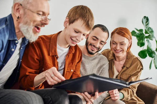 Young gay man looking at photo album near bearded boyfriend and cheerful parents — Stock Photo