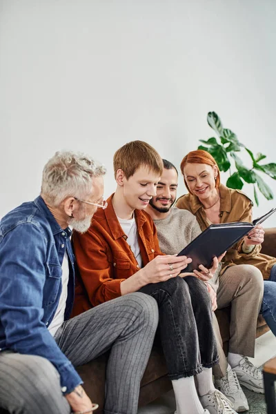 Young gay couple and happy parents sitting on couch in living room and looking at photo album — Stock Photo