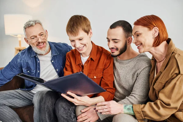 Young and happy gay couple looking at photo album while sitting in living room with pleased parents — Stock Photo