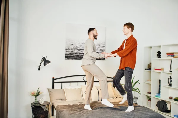Longitud completa de la joven pareja gay cogido de la mano y divertirse en la cama en el dormitorio moderno - foto de stock