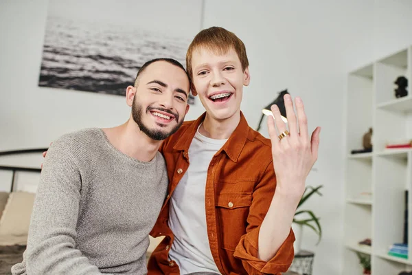 Беззаботный гей показывает обручальное кольцо возле улыбающегося бородатого бойфренда дома — стоковое фото