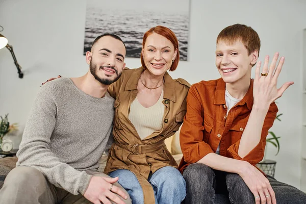 Excited gay man showing wedding ring while smiling at camera with mother and boyfriend at home — Stock Photo
