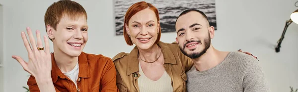 Fröhlicher schwuler Mann zeigt Ehering und lächelt in die Kamera neben Mutter und Freund zu Hause, Banner — Stockfoto