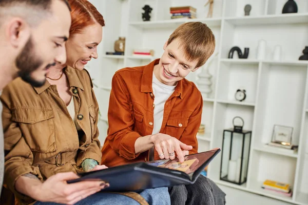 Smiling gay man pointing at photo in album near happy mother and blurred boyfriend at home — Stock Photo