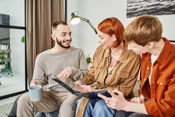 Bearded gay man talking to redhead woman looking at family photo album at home — Stock Photo