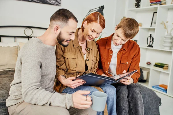 Young gay couple and redhead woman looking at family photo album in bedroom — Stock Photo