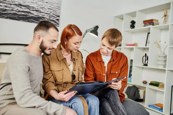 Smiling gay couple sitting in bedroom with mother and looking at family photo album — Stock Photo