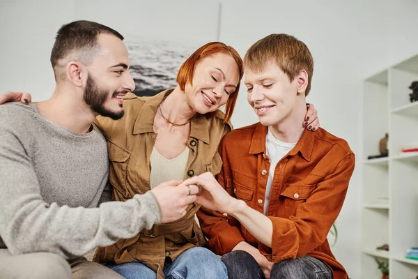 Mujer llena de alegría con los ojos cerrados abrazando hijo con pareja gay en casa - foto de stock