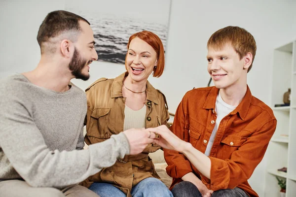 Excited woman laughing near gay man and son showing wedding ring at home — Stock Photo