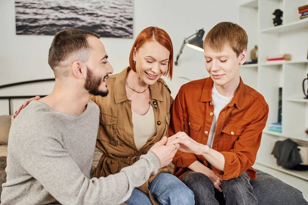 Young gay man showing wedding ring to happy mother near boyfriend at home — Stock Photo