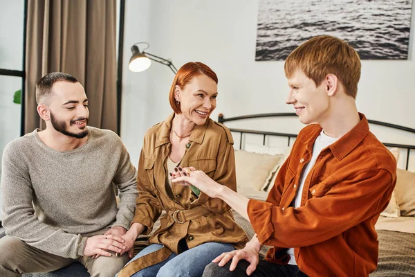 Happy gay man showing wedding ring to pleased mother near boyfriend in bedroom — Stock Photo