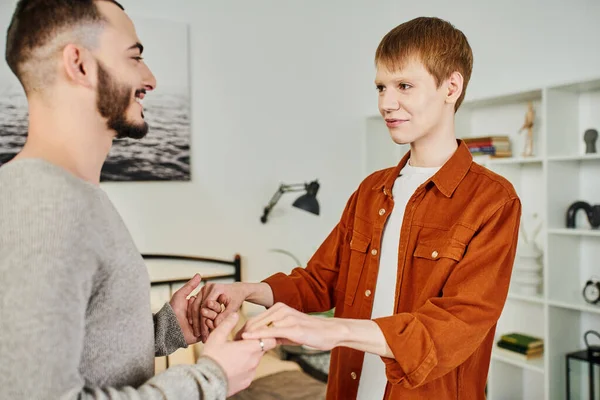 Young and happy gay couple holding hands and looking at each other in bedroom — Stock Photo