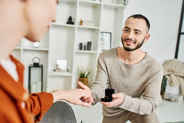 Uomo barbuto sorridente che tiene la mano del fidanzato sfocato mentre fa la proposta di matrimonio a casa — Foto stock