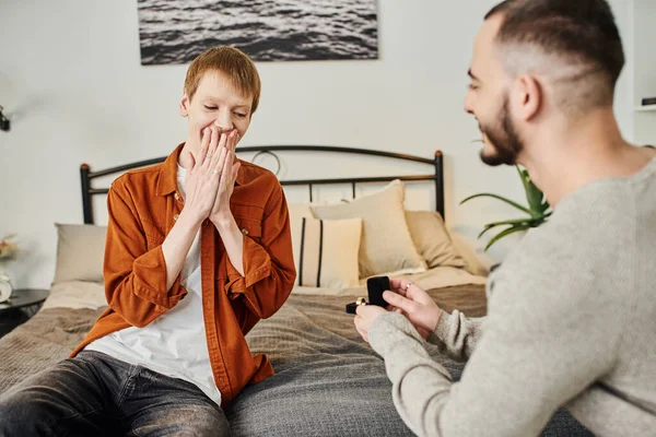 Excited redhead man covering mouth with hands near blurred boyfriend making marriage proposal in bedroom — Stock Photo