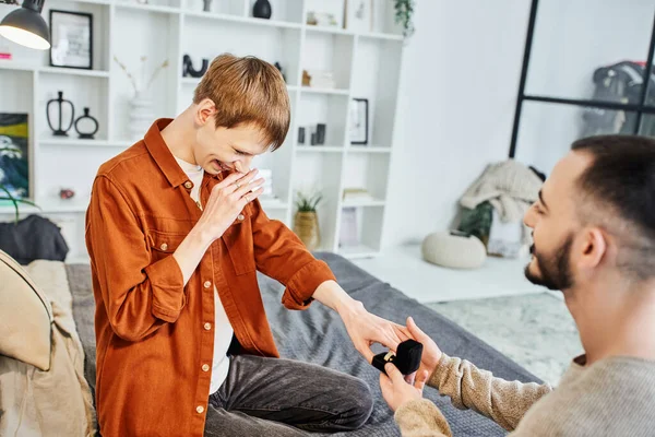 Overjoyed gay man covering mouth with hand near boyfriend making marriage proposal in bedroom — Stock Photo
