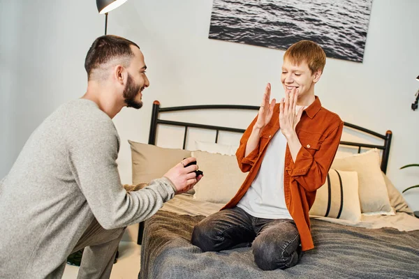 Überglücklicher Rotschopf zeigt Wow-Geste in der Nähe schwuler Partner hält Schmuckschatulle mit Ehering im Schlafzimmer — Stockfoto