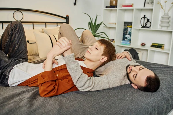 Jóvenes y gays positivos tumbados en el dormitorio moderno y tomados de la mano - foto de stock