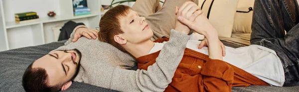 Улыбающийся рыжий мужчина, держащийся за руки с геем, лежа дома на кровати, баннер — стоковое фото