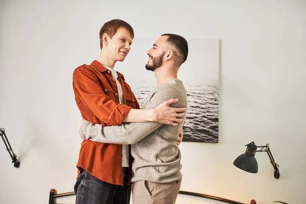 Joven y feliz pareja del mismo sexo tomados de la mano y sonriéndose en casa - foto de stock