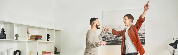 Alegre gay casal segurando mãos enquanto se divertindo no quarto em casa, banner — Fotografia de Stock