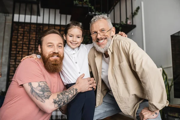 Souriant couple lgbt regardant la caméra et étreignant fille adoptée à la maison — Photo de stock