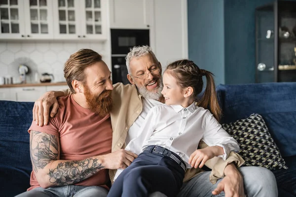 Cheerful gay parents looking at adopted daughter on couch at home — Stock Photo