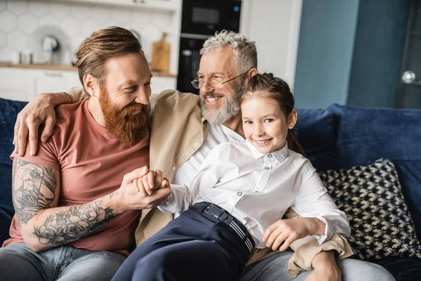 Cheerful gay parents sitting near preteen daughter on couch at home — Stock Photo