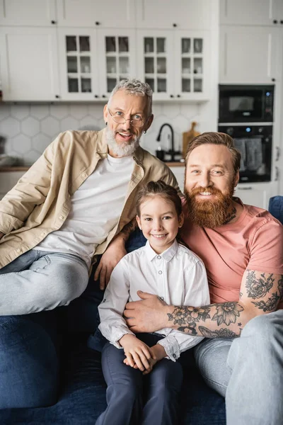 Positive gay parents hugging daughter and looking at camera on couch at home — Stock Photo