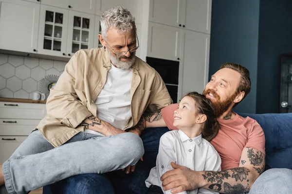 Cheerful and tattooed gay parents sitting near adopted daughter on couch at home — Stock Photo
