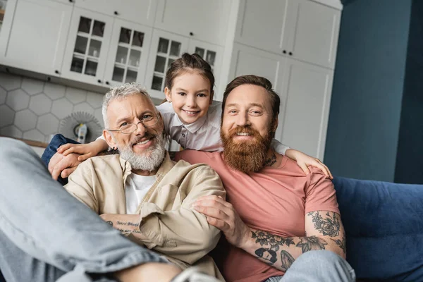 Sorridente ragazza abbracciare tatuato e felice gay padri su divano a casa — Foto stock