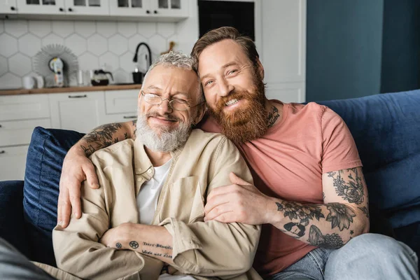 Cheerful gay man hugging bearded partner while sitting on couch at home — Stock Photo