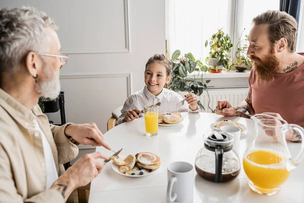 Cheerful preteen girl having pancakes for breakfast near gay parents at home — Stock Photo