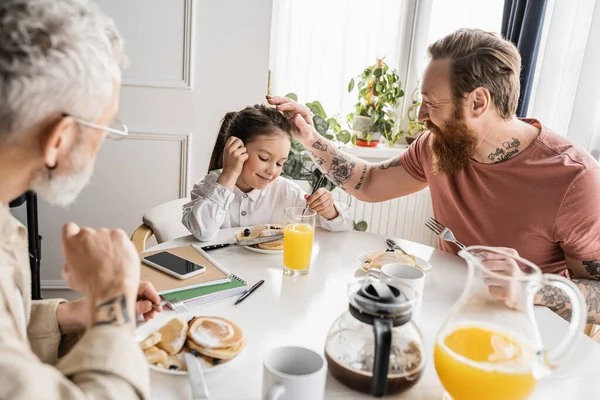 Cheerful gay dad touching head of preteen girl near partner and breakfast at home — Stock Photo