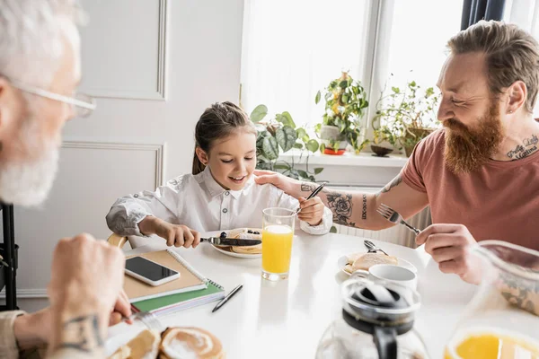 Lächelnder schwuler Vater schaut Tochter beim Pfannkuchenschneiden in der Nähe ihres Partners zu Hause an — Stockfoto