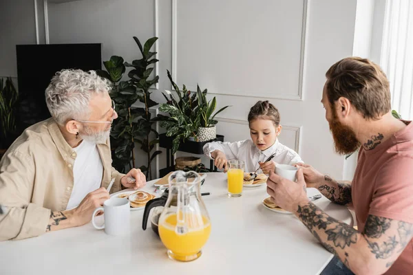 Tattooed gay parents looking at preteen daughter having breakfast at home — Stock Photo