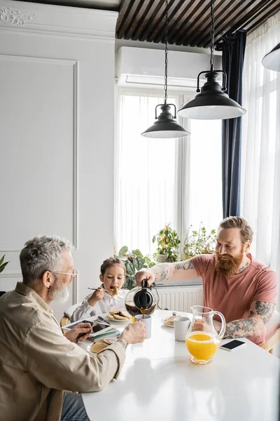 Улыбающийся мужчина наливает кофе рядом с дочерью и гей-партнером во время завтрака дома — стоковое фото