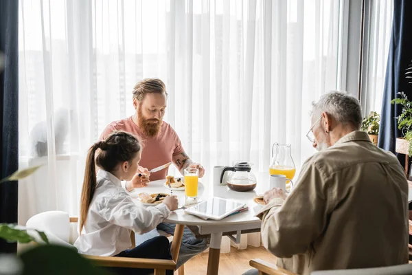 Chica preadolescente desayunando con padres gays cerca de portátiles y tabletas digitales en casa - foto de stock