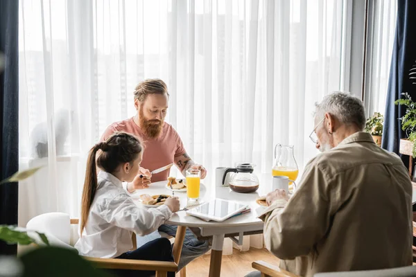 Preteen ragazza avendo colazione con gay genitori vicino notebook e digitale tablet a casa — Foto stock