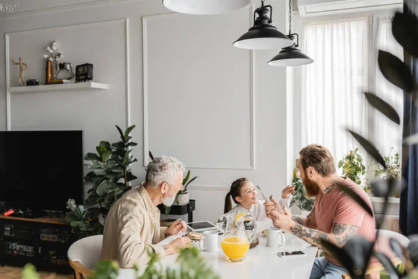 Smiling girl talking to tattooed homosexual parents during breakfast at home — Stock Photo