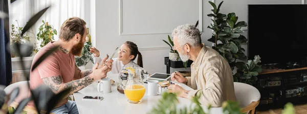 Chica preadolescente positiva hablando con padres homosexuales cerca del desayuno en casa, pancarta - foto de stock