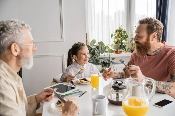 Menina sorridente sentado perto de pais homossexuais e saboroso café da manhã em casa — Fotografia de Stock