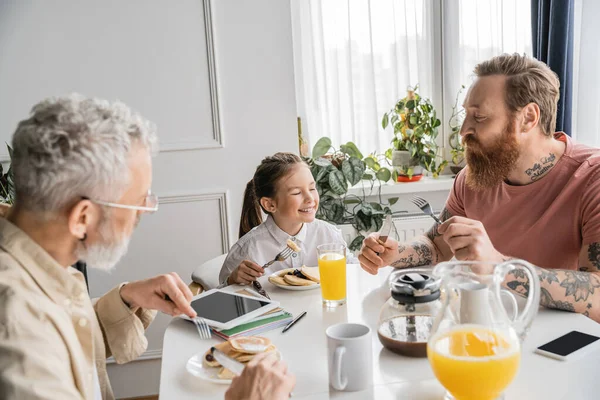 Padres homosexuales mirando a su hija sonriente cerca del desayuno y dispositivos en casa - foto de stock