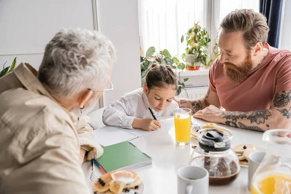 Preteen girl doing homework near gay parents and breakfast at home — Stock Photo