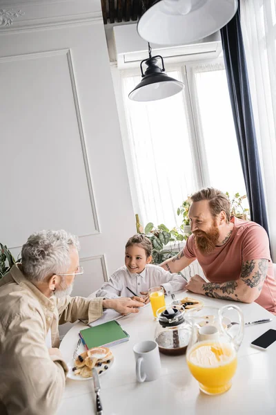 Cheerful kid doing homework near same sex parents during breakfast at home — Stock Photo